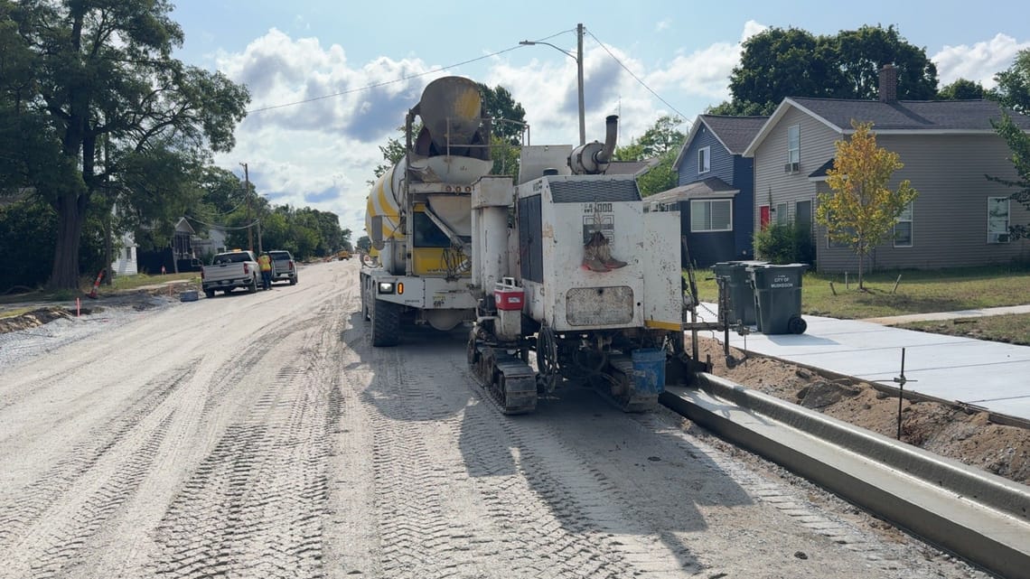 A road construction crew pours a new concrete curb along a residential street. A concrete mixer truck and curb machine are parked on the unpaved roadway, while workers and utility trucks are visible farther down the street. New sidewalks, homes, and City of Muskegon trash carts line the right side under a bright, partly cloudy sky.
