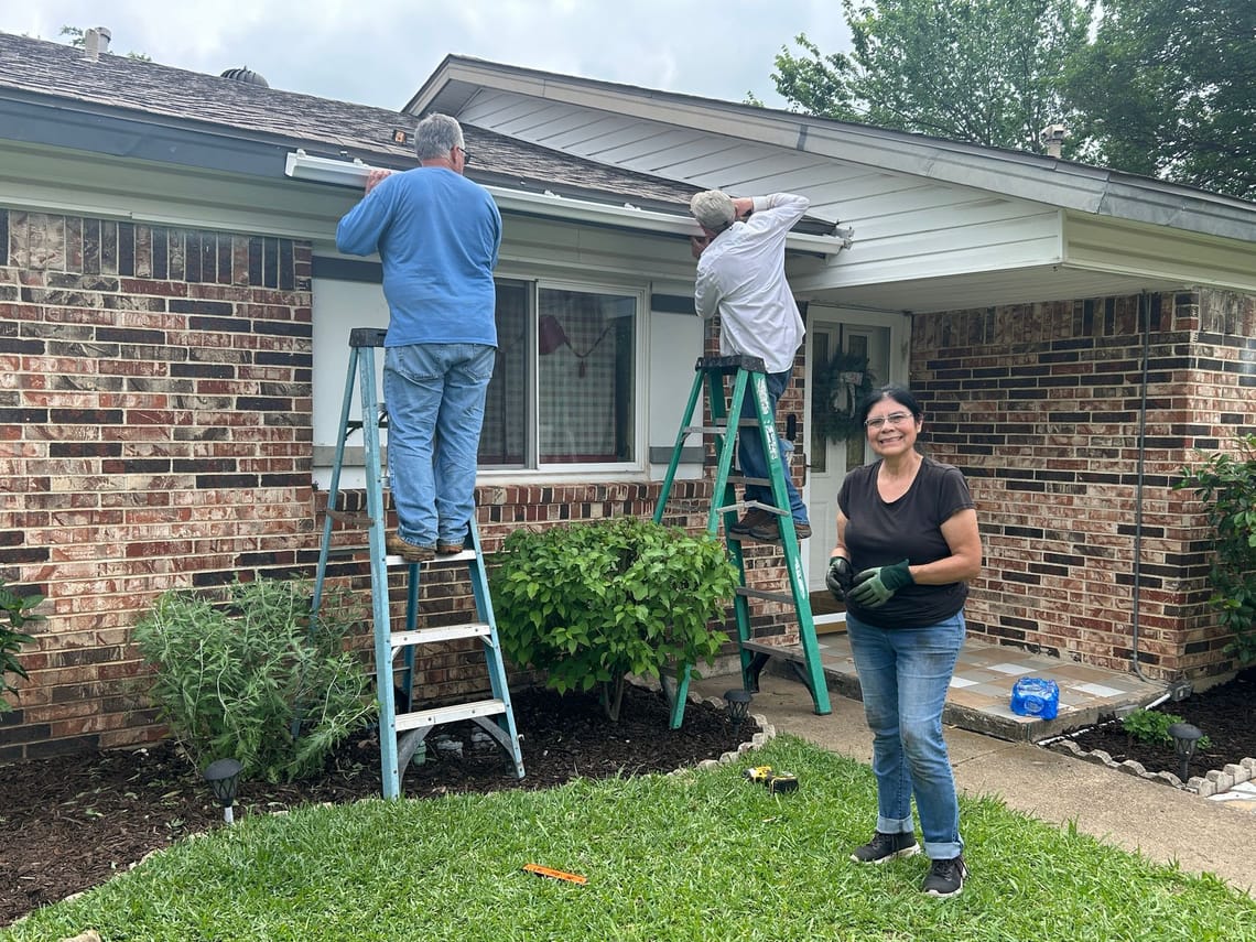 A photo of a woman smiling at the camea wearing work gloves. Behind her, two men stand on ladders attaching a gutter to the front of the house.