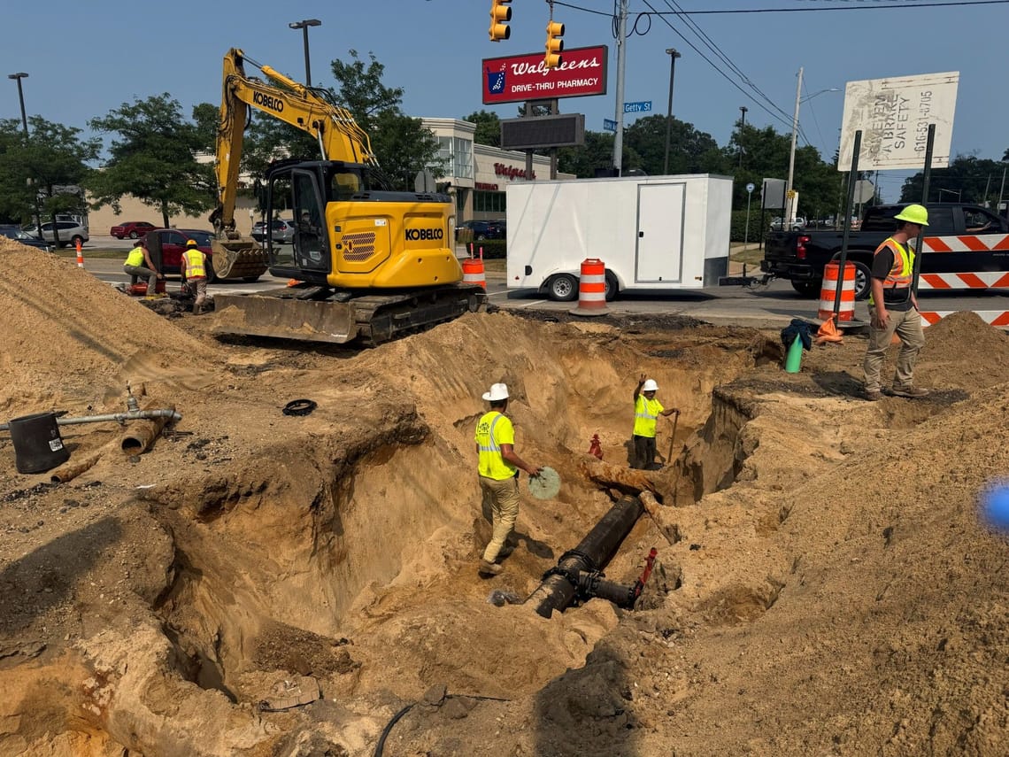 Construction crews install a new water main at a busy intersection. Workers in high-visibility safety gear stand in a large excavation around underground piping while an excavator sits nearby. Traffic control barrels, work vehicles, and a Walgreens store are visible in the background.