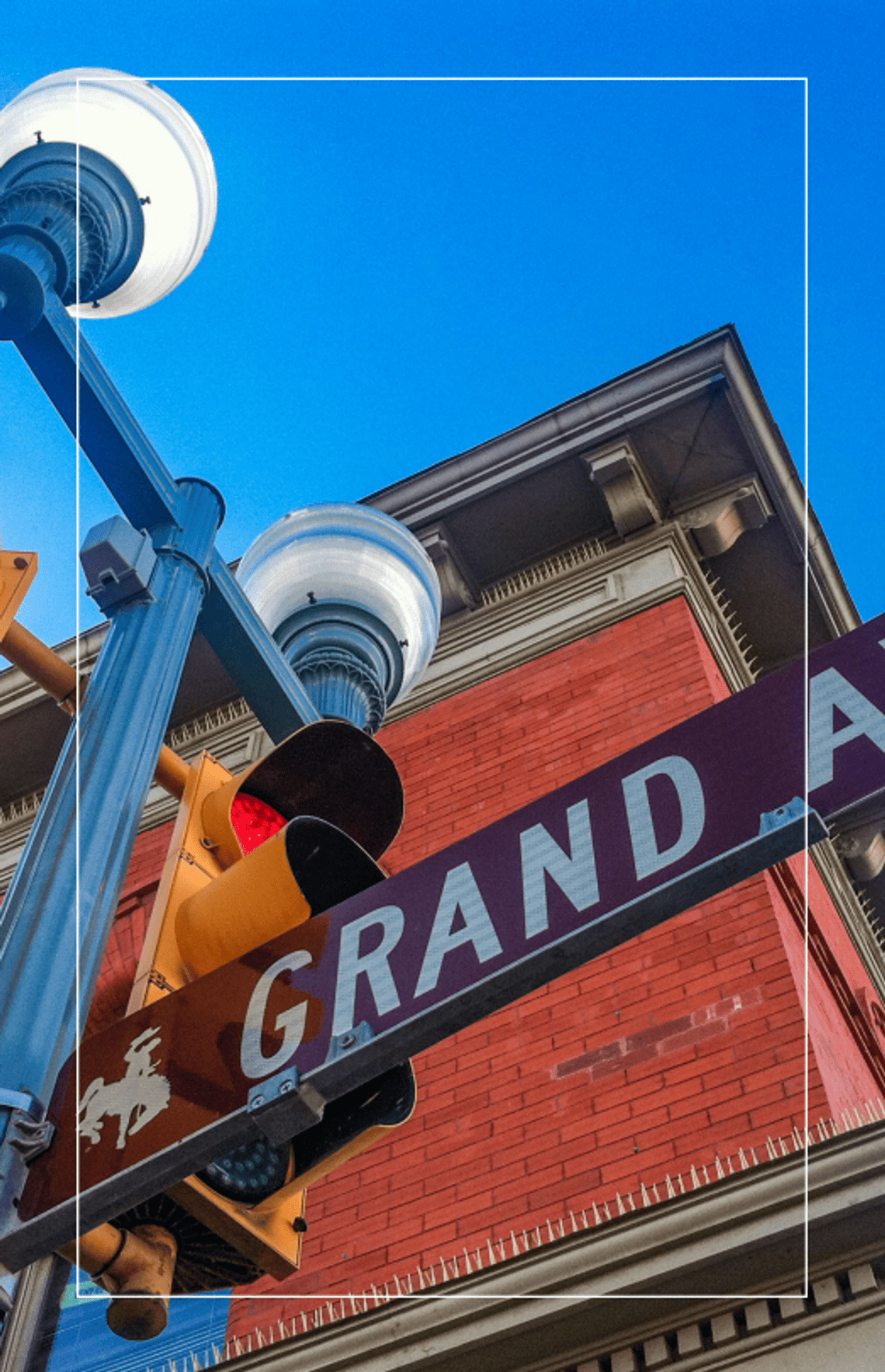 Low-angle view of a Grand Avenue street sign, traffic signal, and decorative streetlights in front of a historic brick building in downtown Laramie.