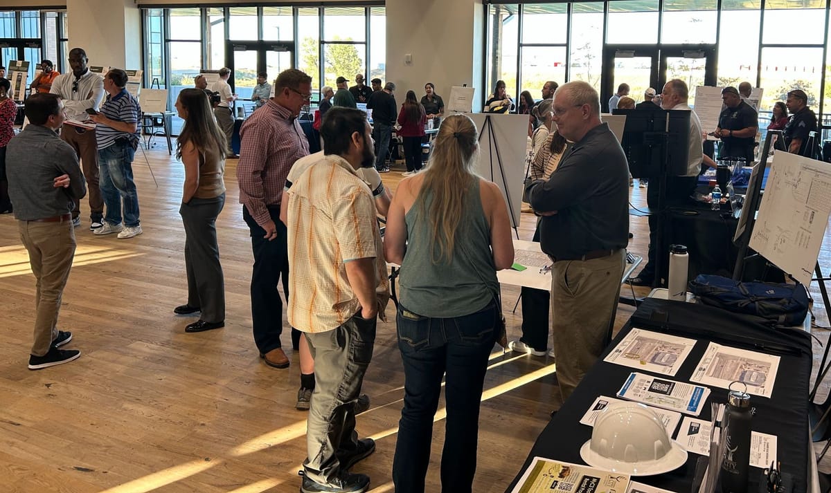 Dozens of people standing in a big room talking at display tables. 