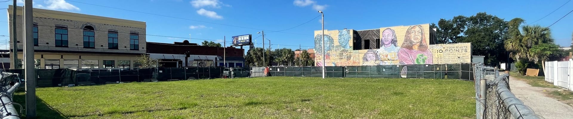 An image showing the future park site located at 2101 W Main St. The site has undergone environmental remediation and has a layer of sod that covers the entire ground surface level. There are two buildings in the background underneath a blue sky with clouds.