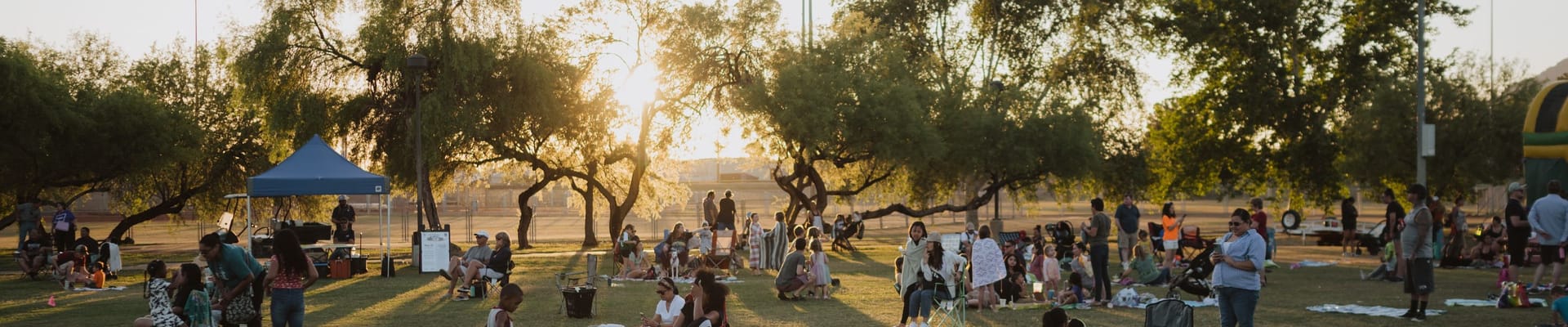 Families and park visitors gather on the grass at Eldorado Park during an outdoor community event, with trees and the setting sun in the background.