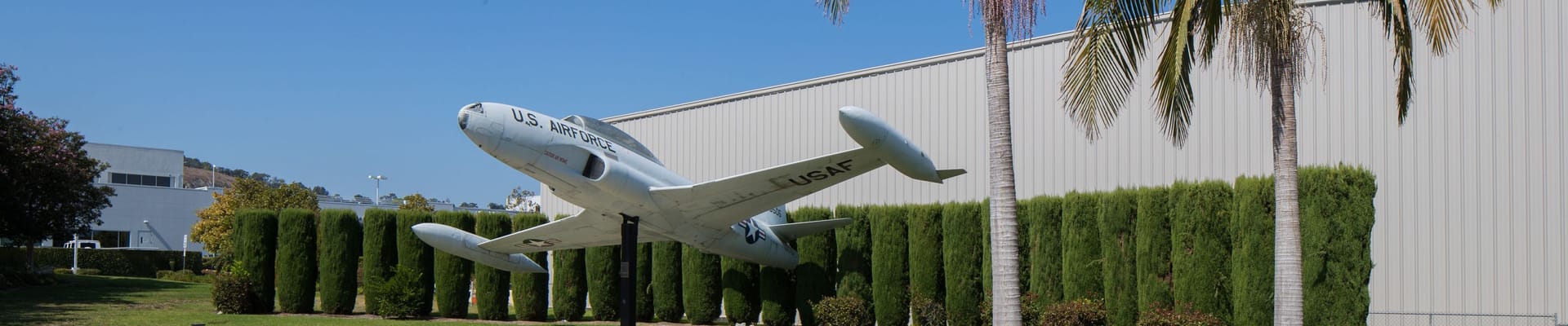 U.S. Air Force jet displayed on a pedestal outside an airport hangar, surrounded by palm trees and landscaping under a clear blue sky.