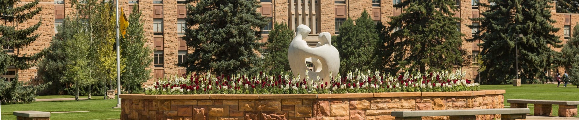 Landscaped plaza with a white abstract sculpture surrounded by flowers, trees, and campus-style brick buildings in the background from the University of Wyoming.