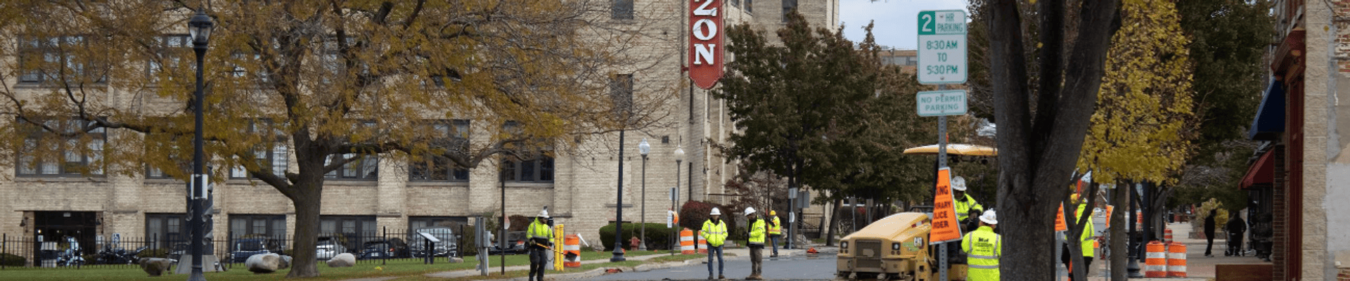 Construction workers resurface a downtown street, with construction equipment, traffic barrels, and a historic building visible along the sidewalk.