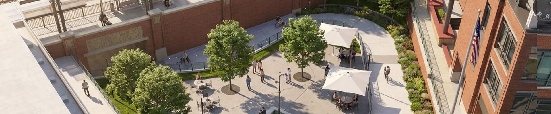 Aerial view of a landscaped public plaza next to a brick building and train station, with trees, seating areas, walkways, ramps and people walking and biking.