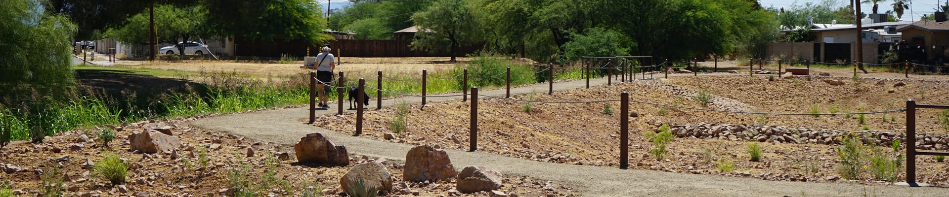 A person walks along a pathway with their black dog. The pathway sits in-between two basins lined with rocks.