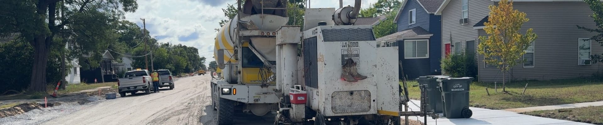 A road construction crew pours a new concrete curb along a residential street. A concrete mixer truck and curb machine are parked on the unpaved roadway, while workers and utility trucks are visible farther down the street. New sidewalks, homes, and City of Muskegon trash carts line the right side under a bright, partly cloudy sky.