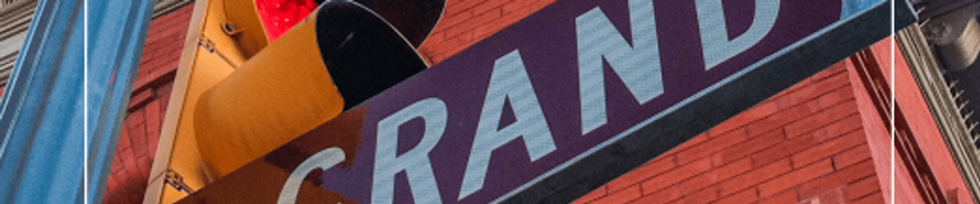Low-angle view of a Grand Avenue street sign, traffic signal, and decorative streetlights in front of a historic brick building in downtown Laramie.