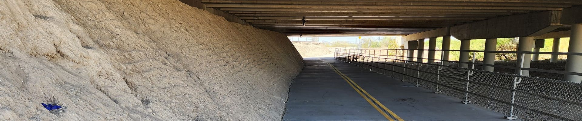 Concrete pathway under a bridge with a dirt embankment, chain-link fencing, support columns, and overhead lighting along the side of the path.