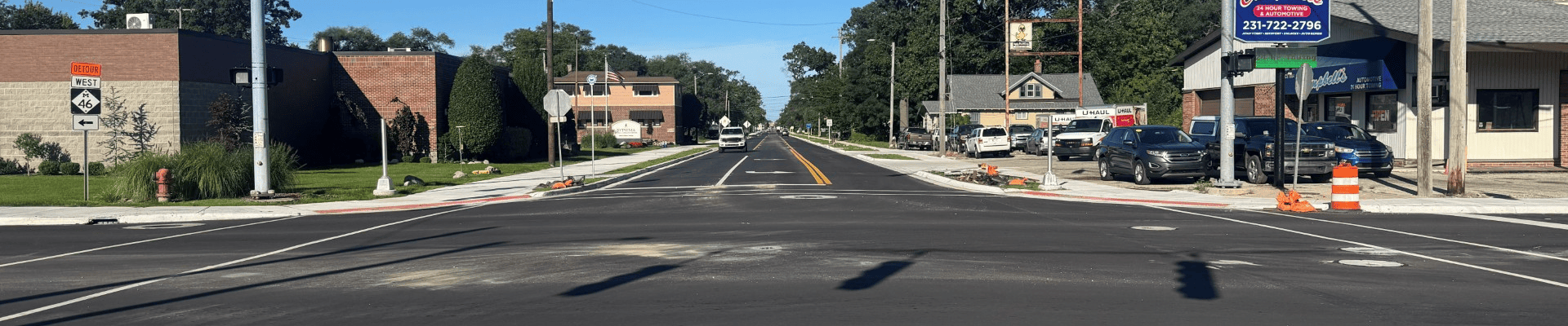 Newly paved roadway at an intersection with traffic signals, fresh lane markings, sidewalks, and construction barrels. Businesses and utility poles line both sides of the street under a clear blue sky.