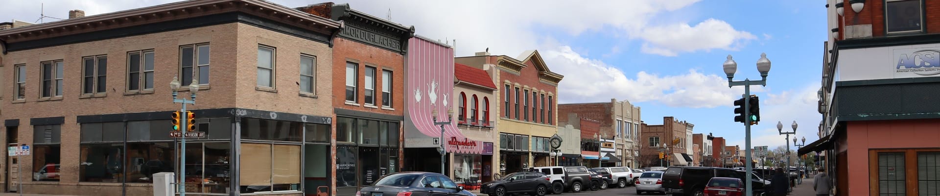 Downtown Laramie streetscape with historic storefronts, parked cars, and decorative streetlights under a partly cloudy sky.