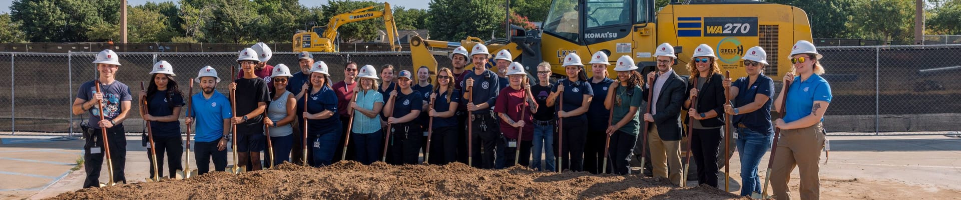 people stand with shovels in dirt with excavator in background.