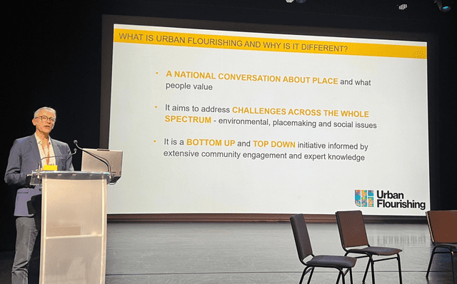 Hugo Nowell standing at a podium next to a big screen displaying an overview of what Urban Flourishing is