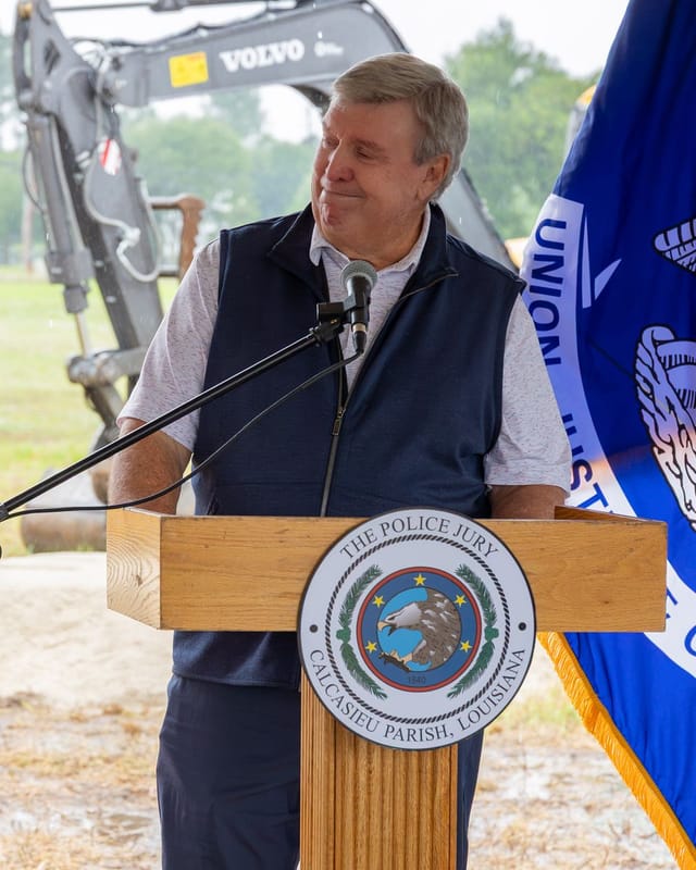 Officials speaking at podium with flags and an excavator in the background