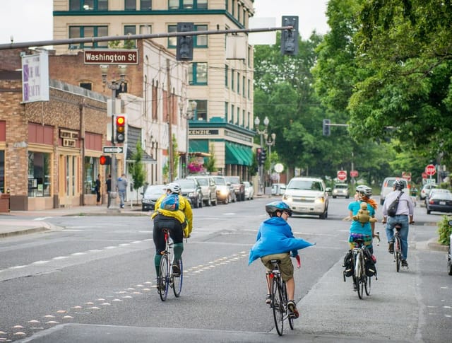 Cyclists ride eastbound on 6th Street, crossing Washington Street on an overcast day. Cars, large trees, and storefronts appear in the background. . 