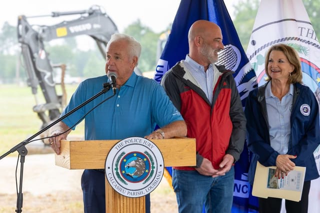 Officials speaking at podium with flags and an excavator in the background