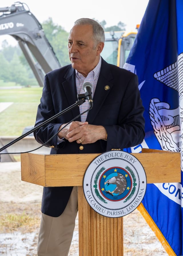 Officials speaking at podium with flags and an excavator in the background