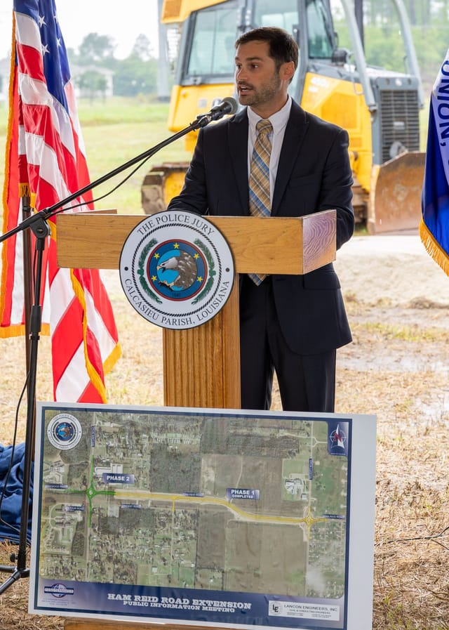 Officials speaking at podium with flags and an excavator in the background