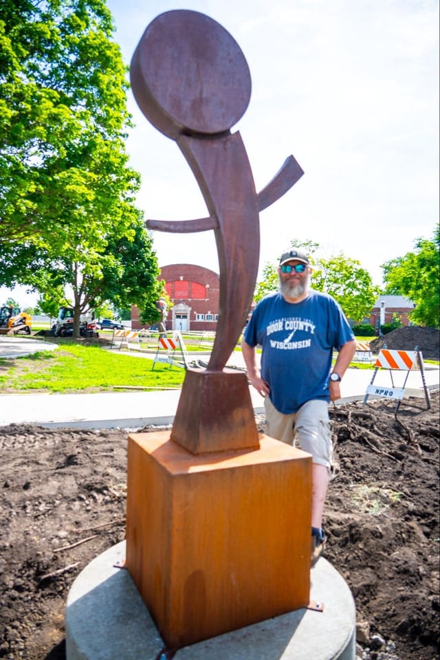The artist next to a 6’ tall carbon steel sculpture on a 3’ base, the gleeful piece mimics an enthusiastic person with raised arms