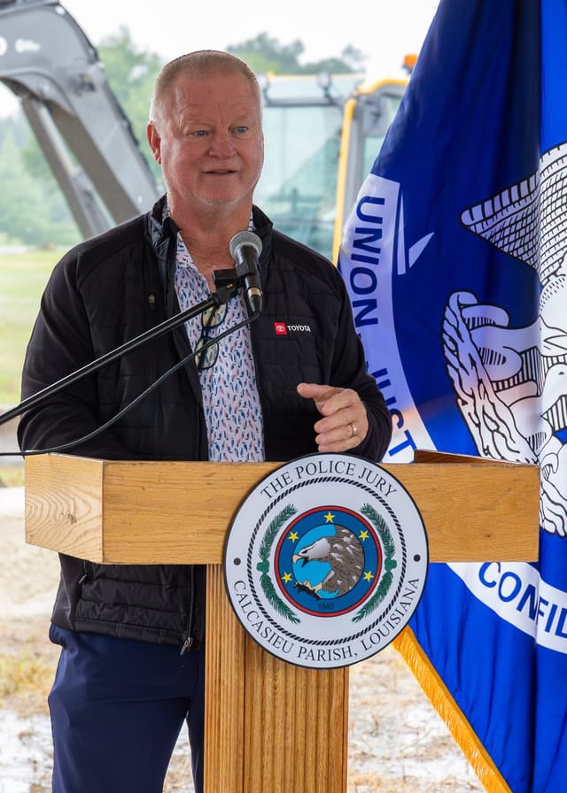 Officials speaking at podium with flags and an excavator in the background