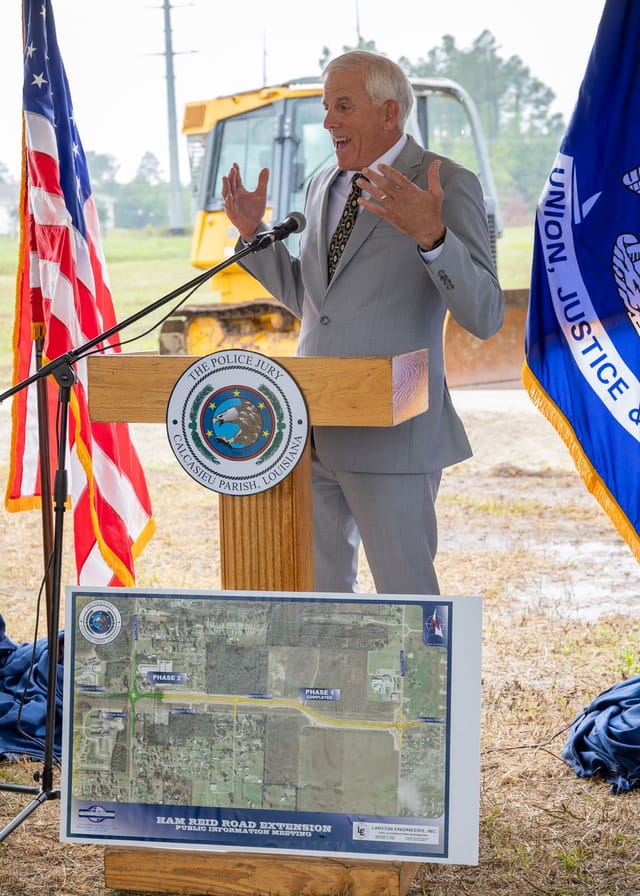 Officials speaking at podium with flags and an excavator in the background