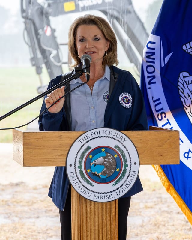 Officials speaking at podium with flags and an excavator in the background