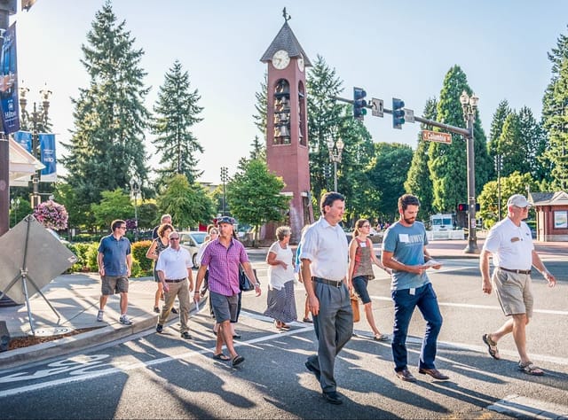 A group of pedestrians walk across Columbia street near Esther Short Park. The Salmon Run Bell Tower is visible behind the pedestrians, surrounded by tall trees and street signs. People of various ages walk together, casting long shadows on the crosswalk.