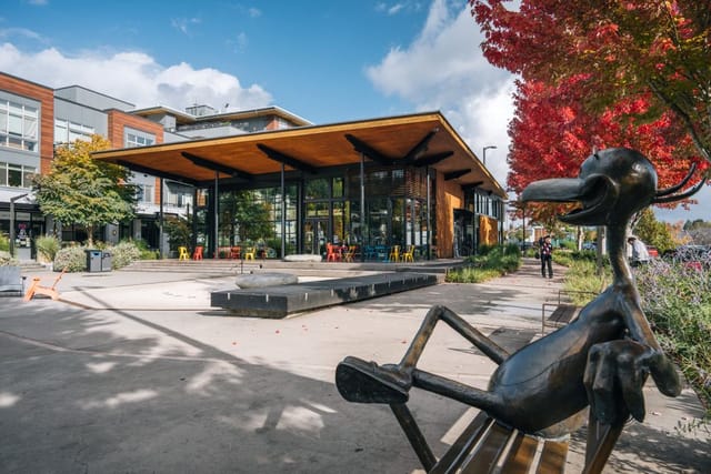 Open plaza with a bronze heron sculpture sitting on it with a fall foliage tree in the background a large building with large windows. 