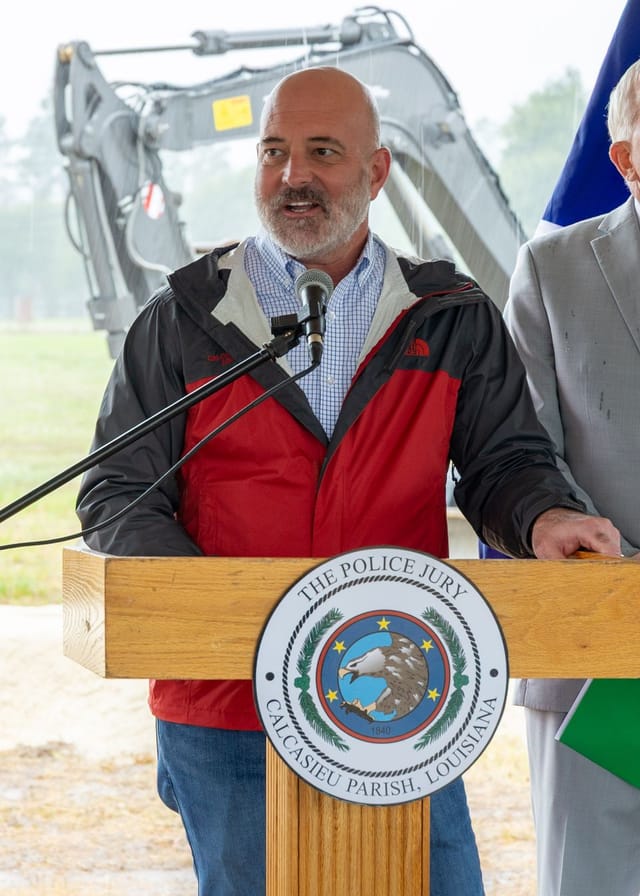 Officials speaking at podium with flags and an excavator in the background