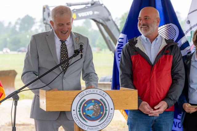 Officials speaking at podium with flags and an excavator in the background