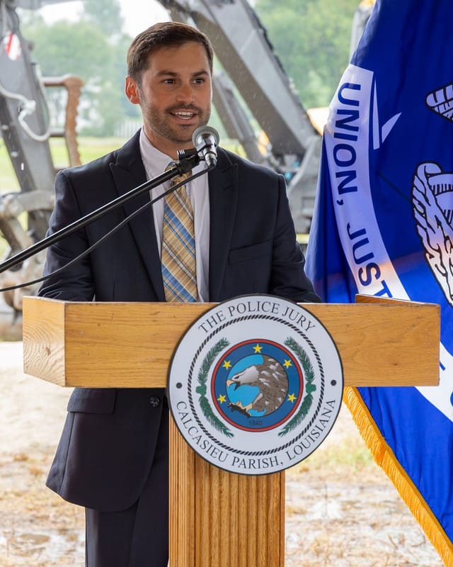 Officials speaking at podium with flags and an excavator in the background