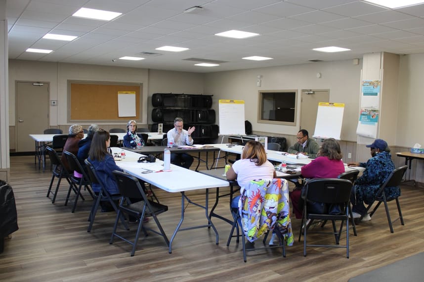 A group of community members sit around tables during a community planning discussion, with posters and easels displaying notes around the room.