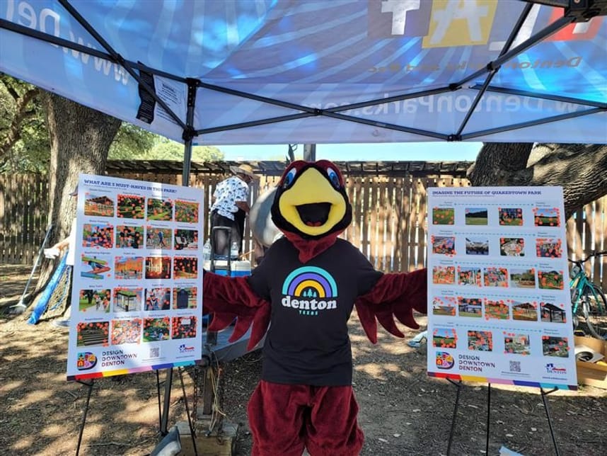 Reddy the Parks and Recreation mascot with a black shirt that says denton Texas, standing between two poster boards under a tent