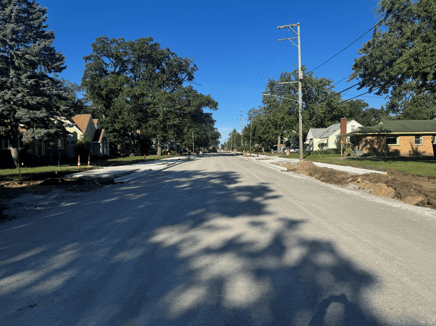 Residential street under construction with a gravel road base, newly poured concrete curbs and sidewalks, exposed soil, and orange construction barrels. Houses, mature trees, and utility poles line both sides of the street under a clear blue sky.