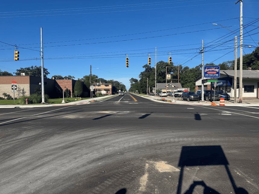 Newly paved roadway at an intersection with traffic signals, fresh lane markings, sidewalks, and construction barrels. Businesses and utility poles line both sides of the street under a clear blue sky.