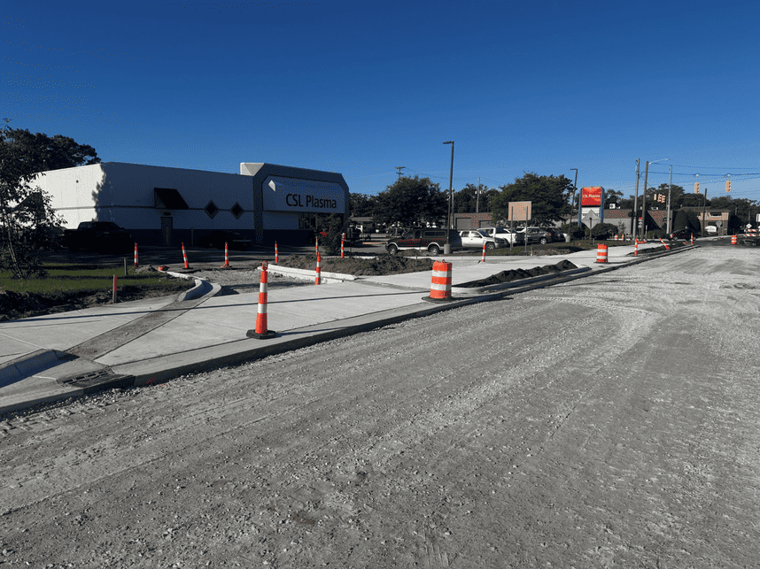 Road construction underway in front of a CSL Plasma building, with newly poured concrete curbs and sidewalks, exposed soil, gravel roadway, and orange construction barrels marking the work area.