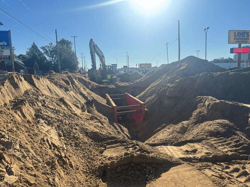 Construction site showing a deep trench with a red trench box, large piles of excavated soil, and an excavator and worker in the background. Businesses and utility poles line the roadway, with a clear blue sky overhead.