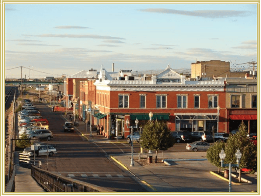 View of historic downtown Laramie with brick storefronts, parked cars, street trees, and decorative lighting along a commercial street.