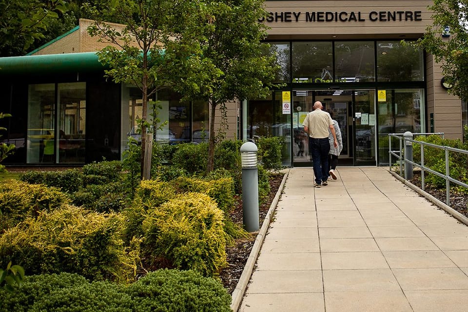 Image shows a man and a woman walking along a paved path towards the entrance to Bushey Medical Centre, The pathh is edged with greenery on one side and a railing on the other.