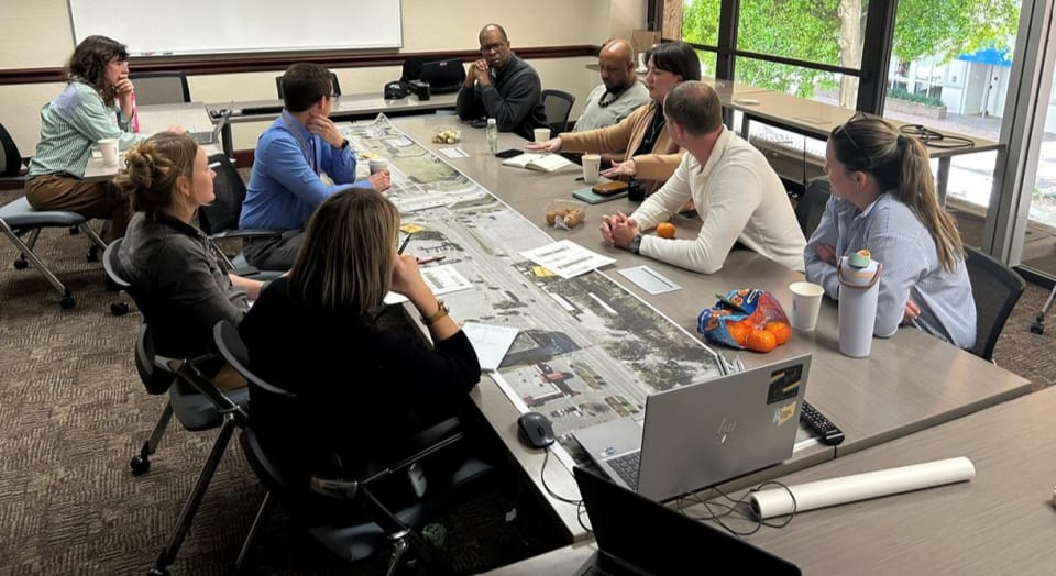 People gathered and engaging in conversation in a conference room around a long table with a roll plot showing a satellite view of Bird St.