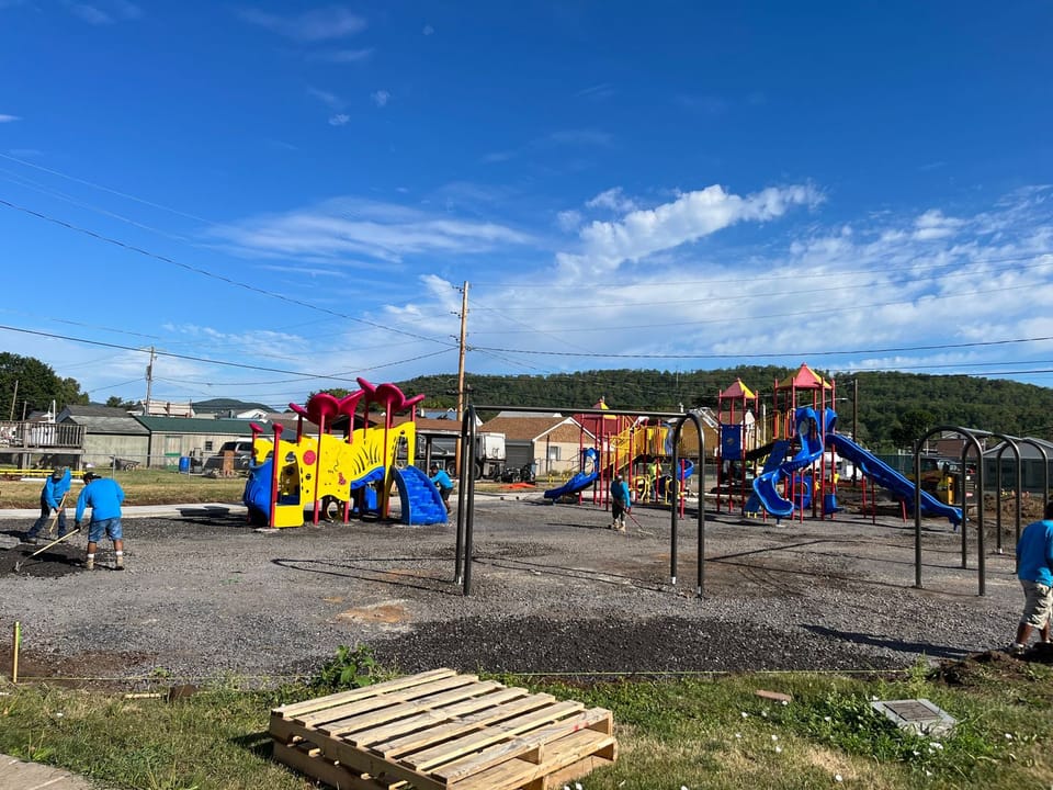 Image showing the continued construction at Piper-Harmon Playground. The installed swing sets and play structures are shown with a gravel surface under them as crews rake and stamp the surface to prepare to install rubber fall surface.