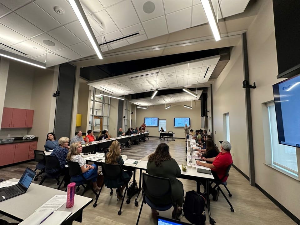 Team members sitting at tables made in a U shape looking at two tvs with three instructors talking