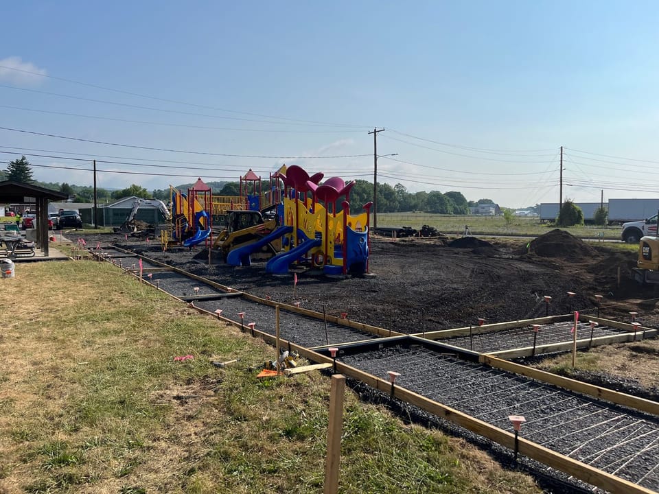 Image showing the progress of construction at the Piper-Harmon Playground. A framed-out walkway ready for concrete to be poured can be seen in the foreground. Exposed gravel can also be seen in the background of the image under the existing play equipment as it is prepared for rubber fall-safe surface.