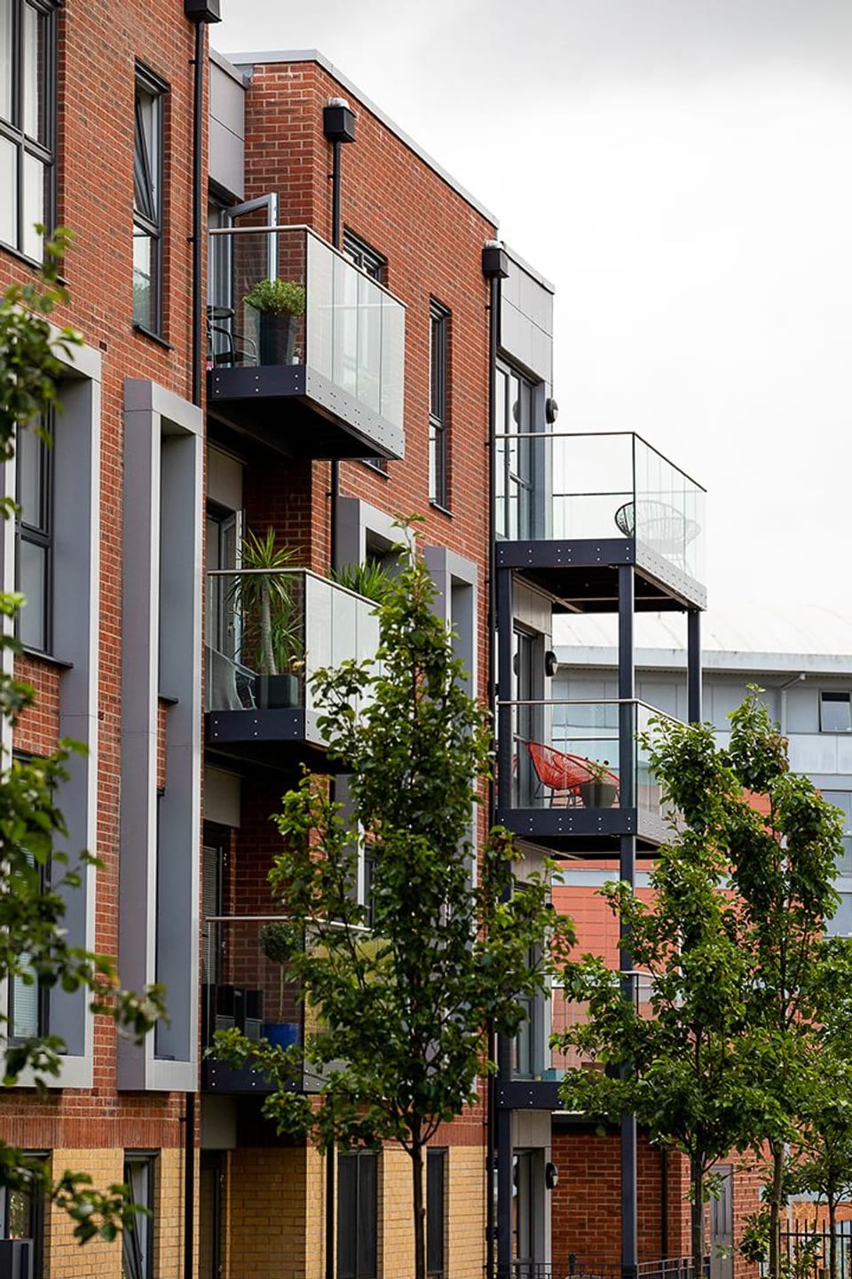 Image shows a photograph of modern flats in Borehamwood. The building is of red and yellow brick with balconies, and small street trees in the foreground.
