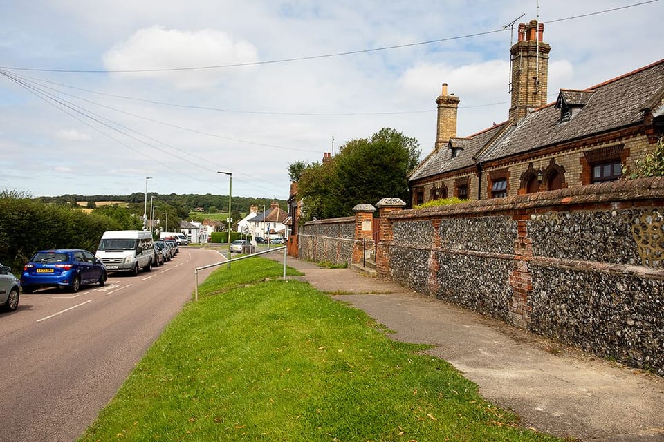 A photograph of South Mimms Village, showing a road with parked cars, a grass verge and a flint wall and domestic buildings